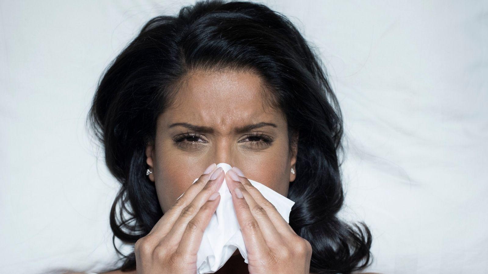 image of woman with dark shoulder length hair sneezing into a white tissue