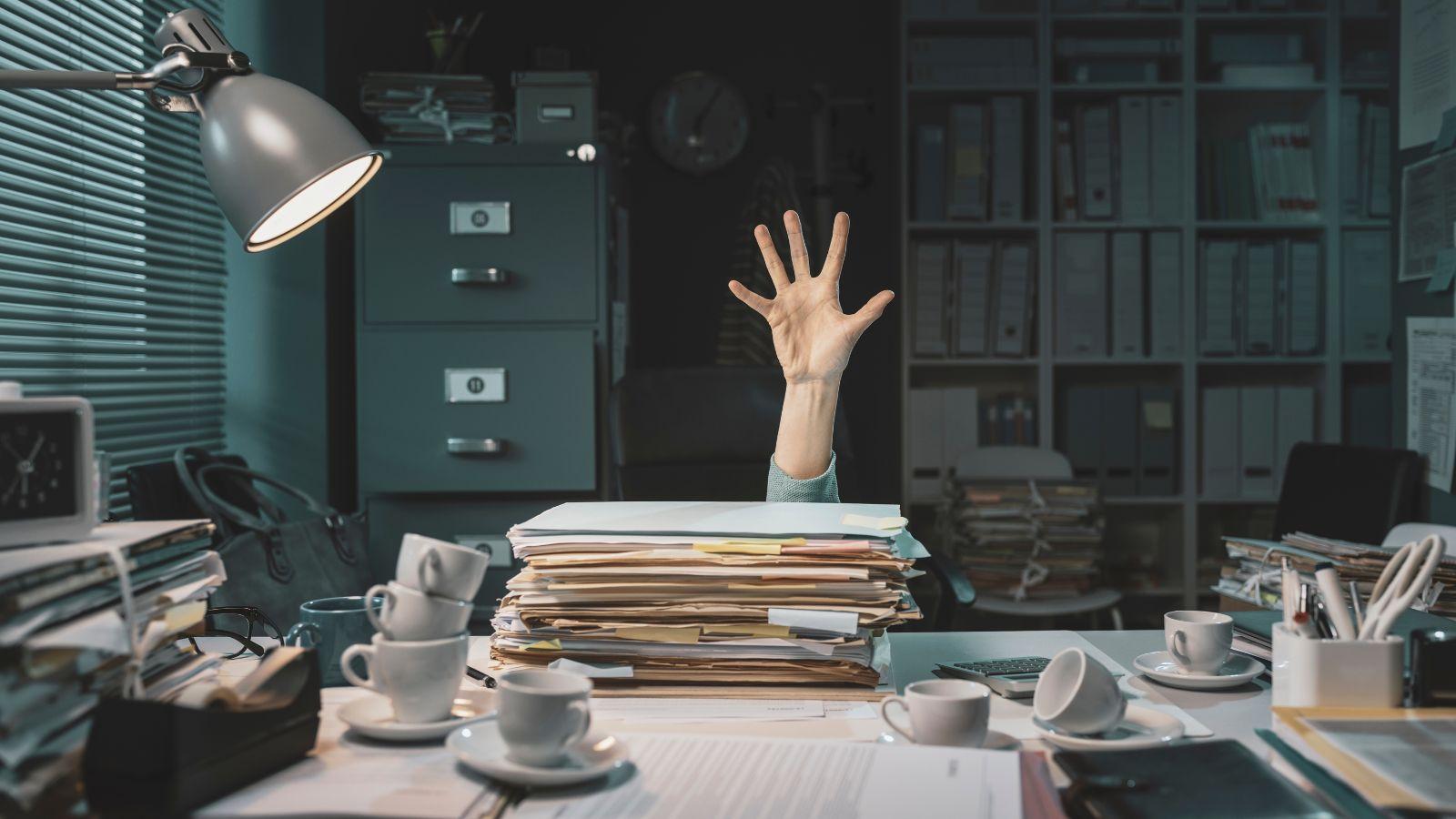 overcome stress. Image of a pile of paperwork on a desk with a hand waving for help