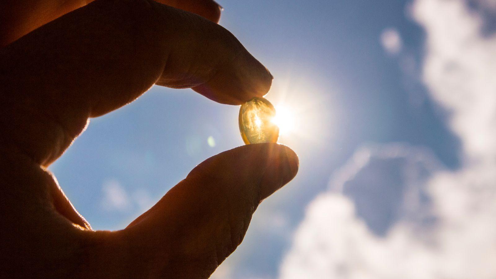 autumn self care tips vitamin d. Image of vitamin d capsule being held up to the sunlight. With clouds in the background and a blue sky