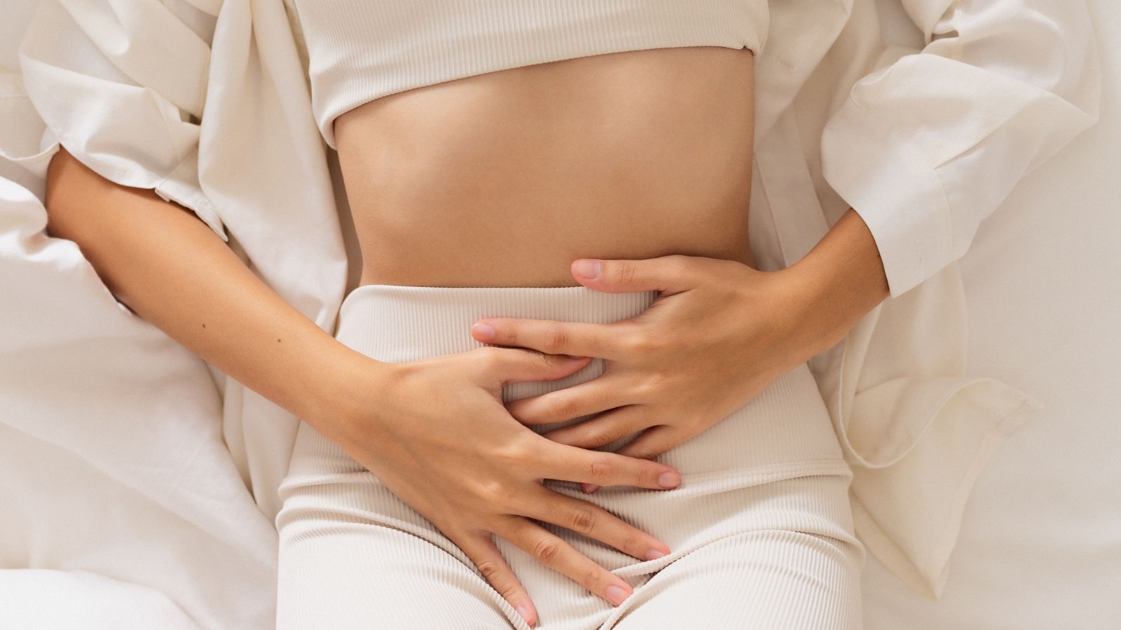 Uses and benefits of rose essential oil. Helps womens problems. Image of young woman wearing a light coloured pair of trousers, clutching her stomach region