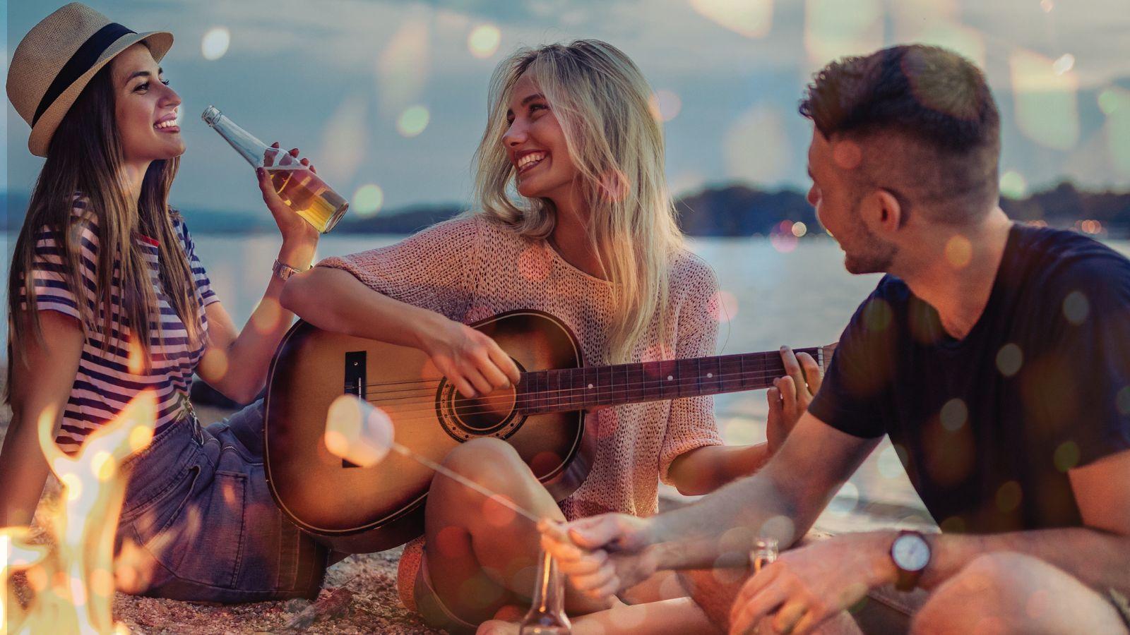 Image of a group of friends making smores around a campfire whilst someone plays a guitar. Summer vibes