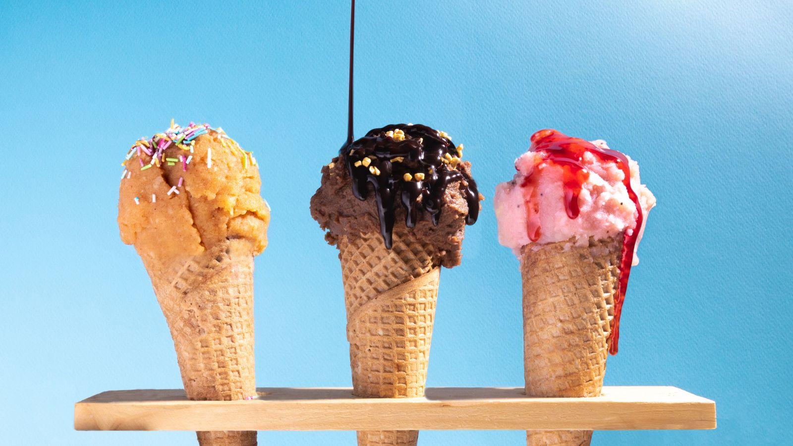 image of three ice creams in cones on a sky blue background, the cones appear to be honey comb, chocolate and raspberry ripple ice cream
