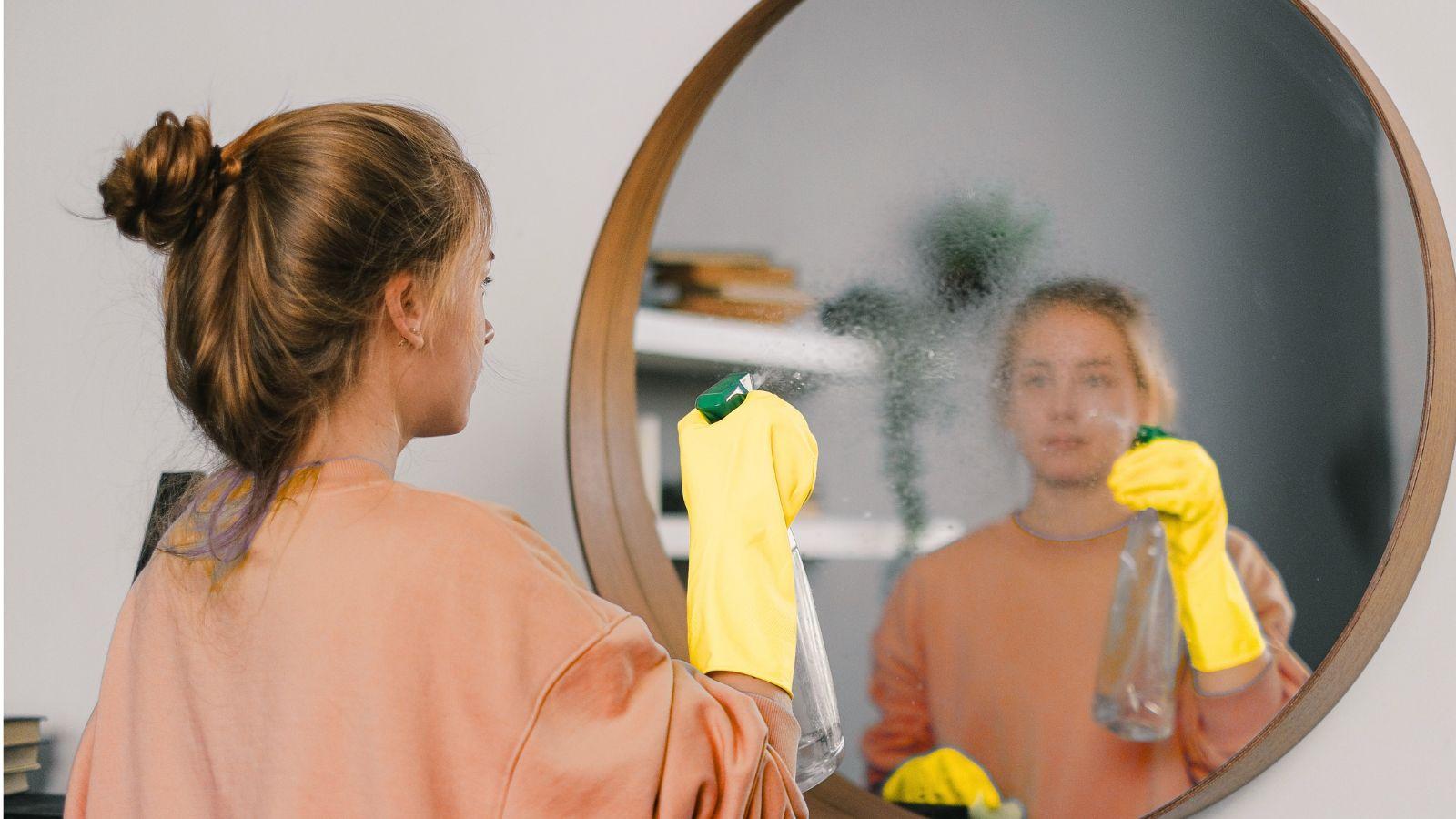 Raise your home's vibration by polishing mirrors, removing cobwebs Image of a woman in yellow rubber gloves polishing a mirror